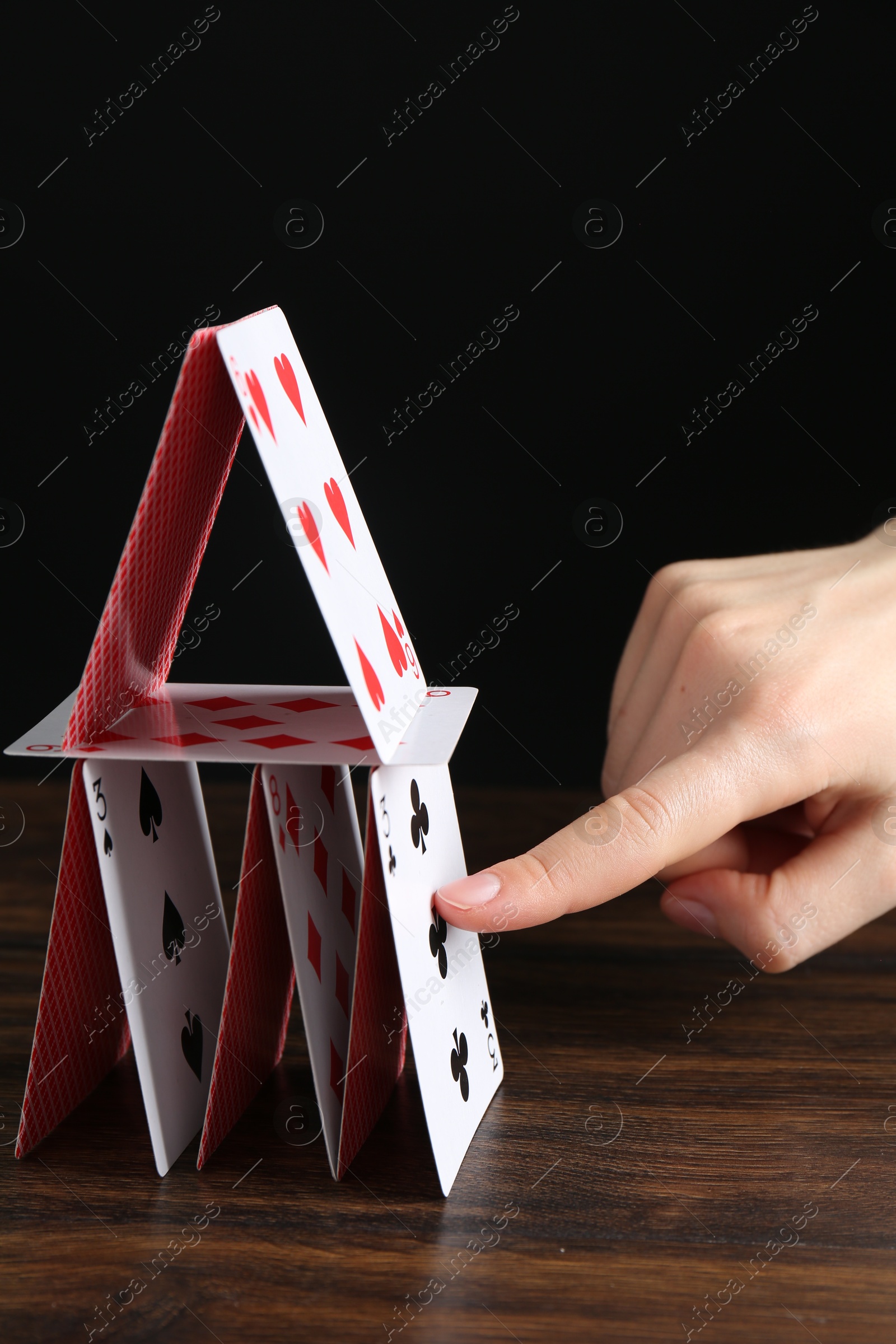 Photo of Woman destroying house of playing cards at wooden table against black background, closeup