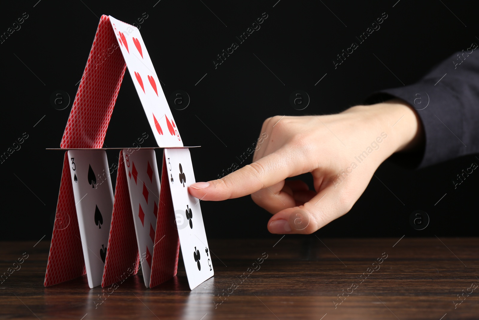 Photo of Woman destroying house of playing cards at wooden table against black background, closeup