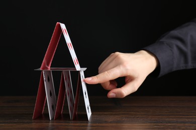 Woman destroying house of playing cards at wooden table against black background, closeup