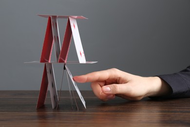 Photo of Woman destroying house of playing cards at wooden table against grey background, closeup