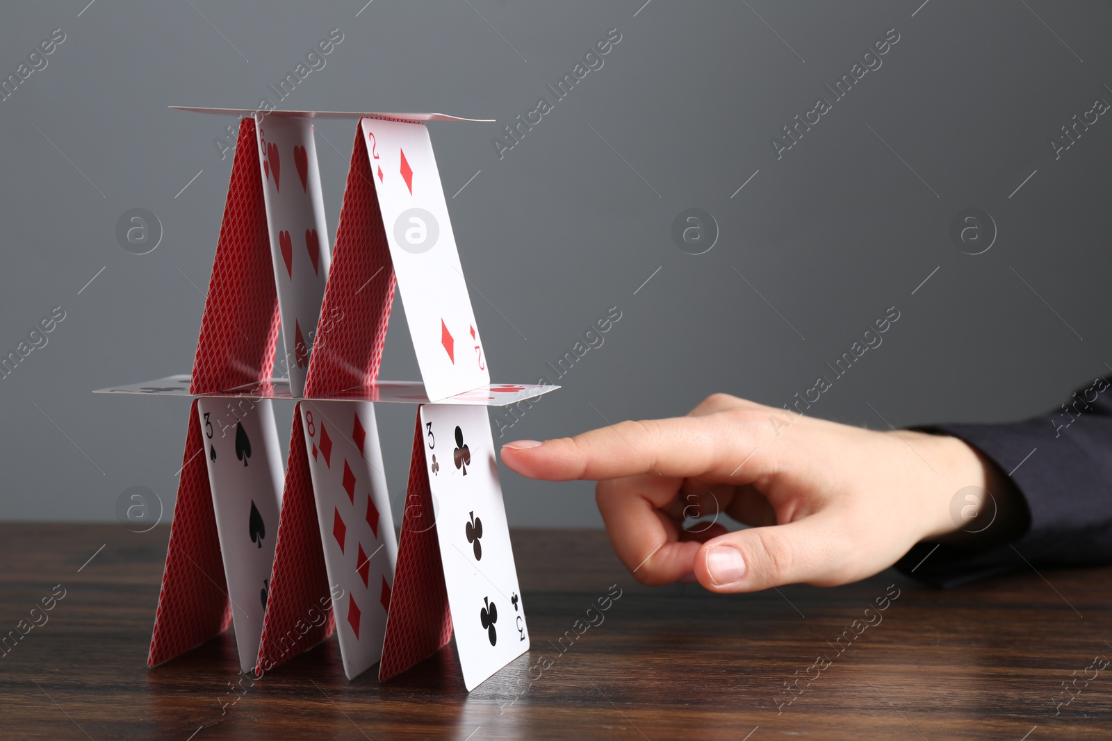 Photo of Woman destroying house of playing cards at wooden table against grey background, closeup