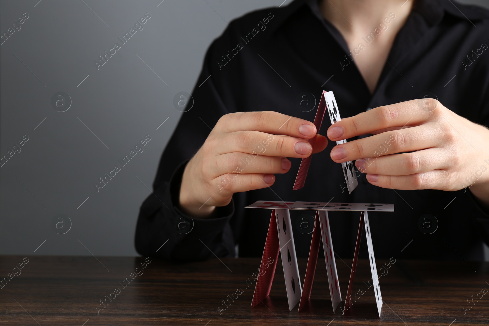 Photo of Woman building house of playing cards at wooden table against grey background, closeup. Space for text