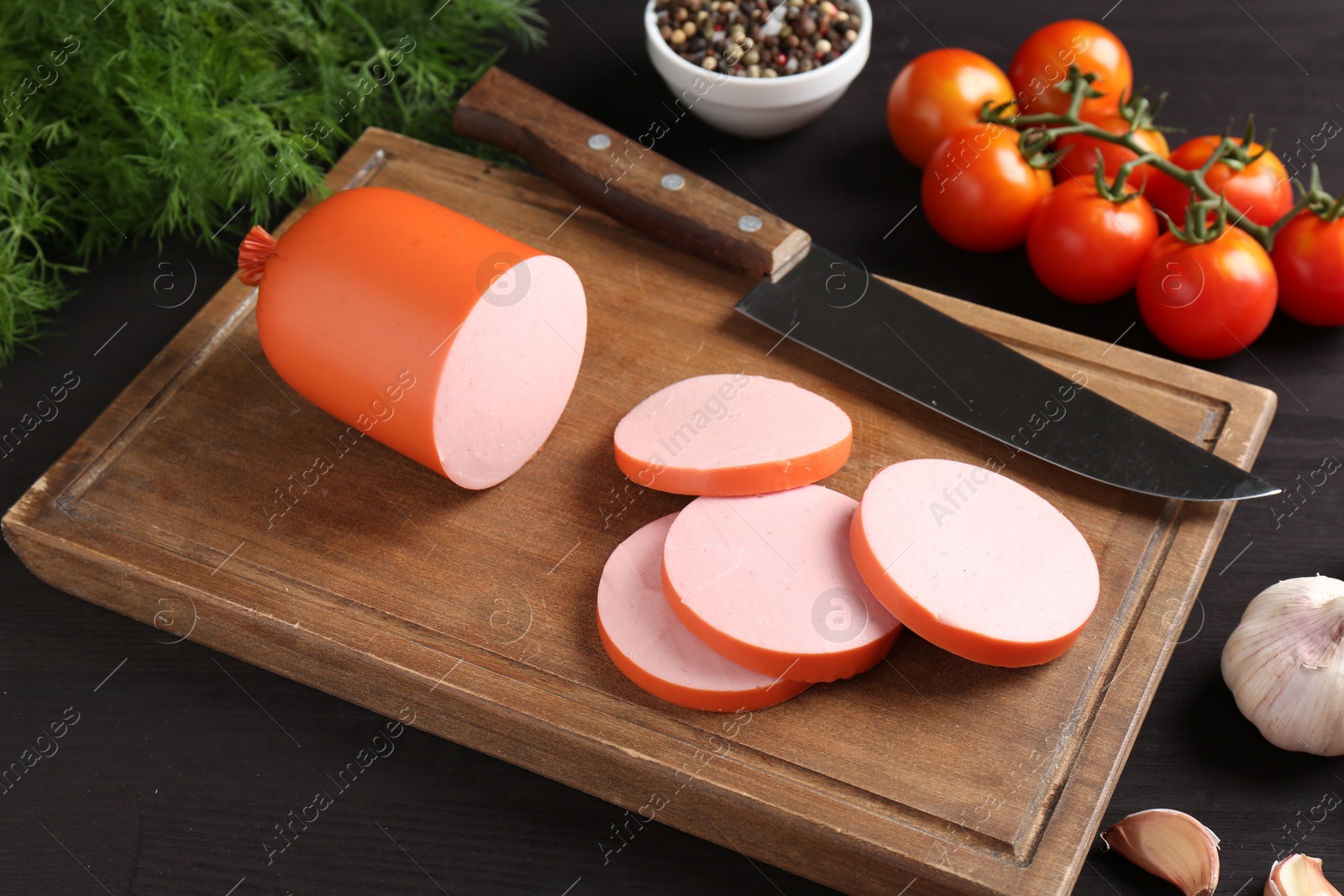 Photo of Tasty boiled sausage, knife, spices and tomatoes on black wooden table