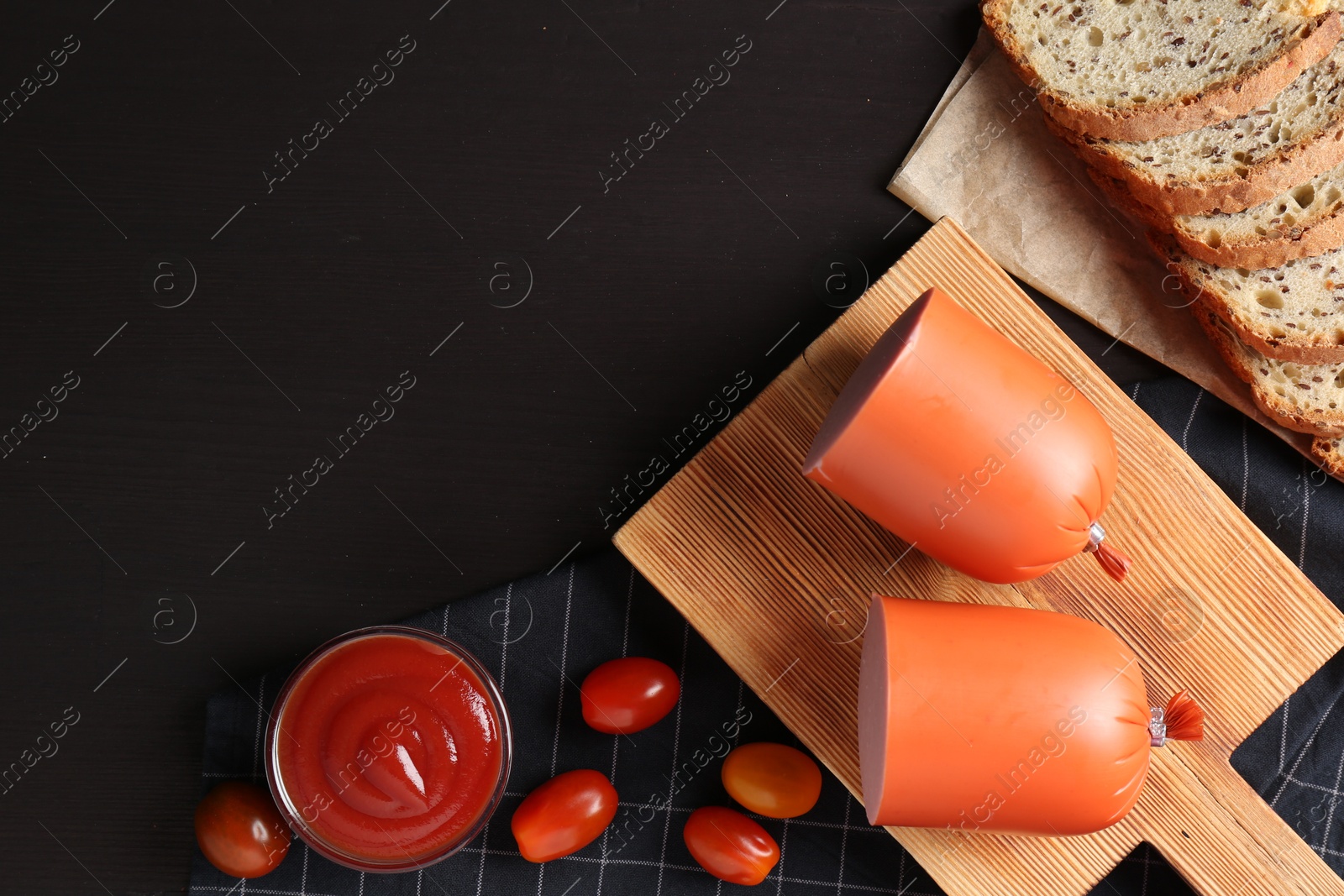 Photo of Tasty boiled sausage, ketchup, tomatoes and bread on black wooden table, top view. Space for text