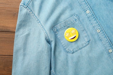 Image of Badge with star-eyes face attached to pocket of denim shirt on wooden background, top view