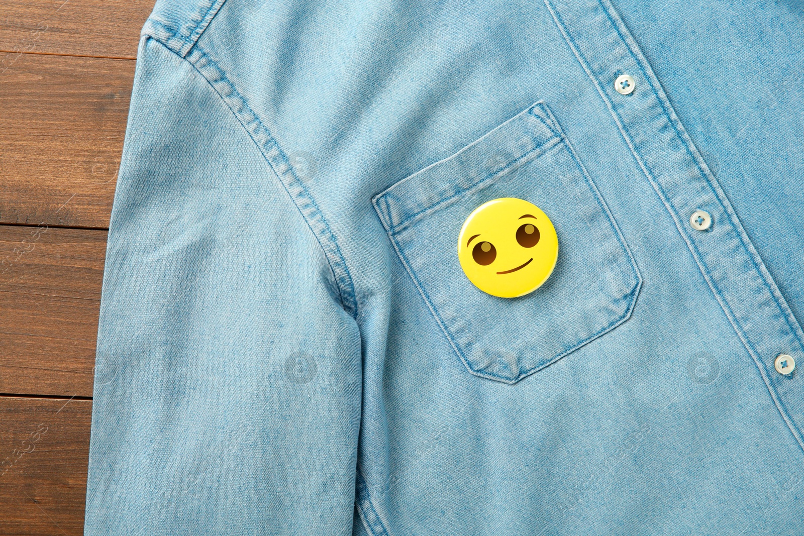 Image of Badge with smiling face attached to pocket of denim shirt on wooden background, top view