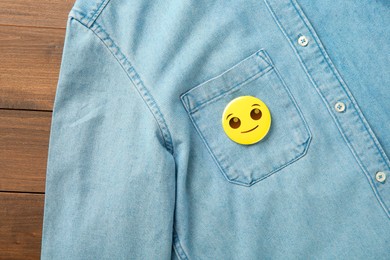 Badge with smiling face attached to pocket of denim shirt on wooden background, top view