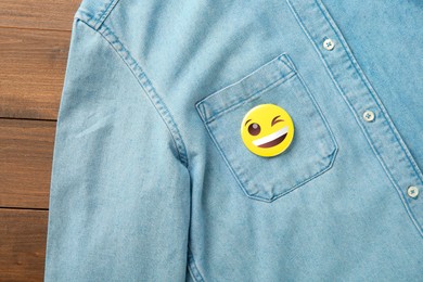 Image of Badge with winking face attached to pocket of denim shirt on wooden background, top view