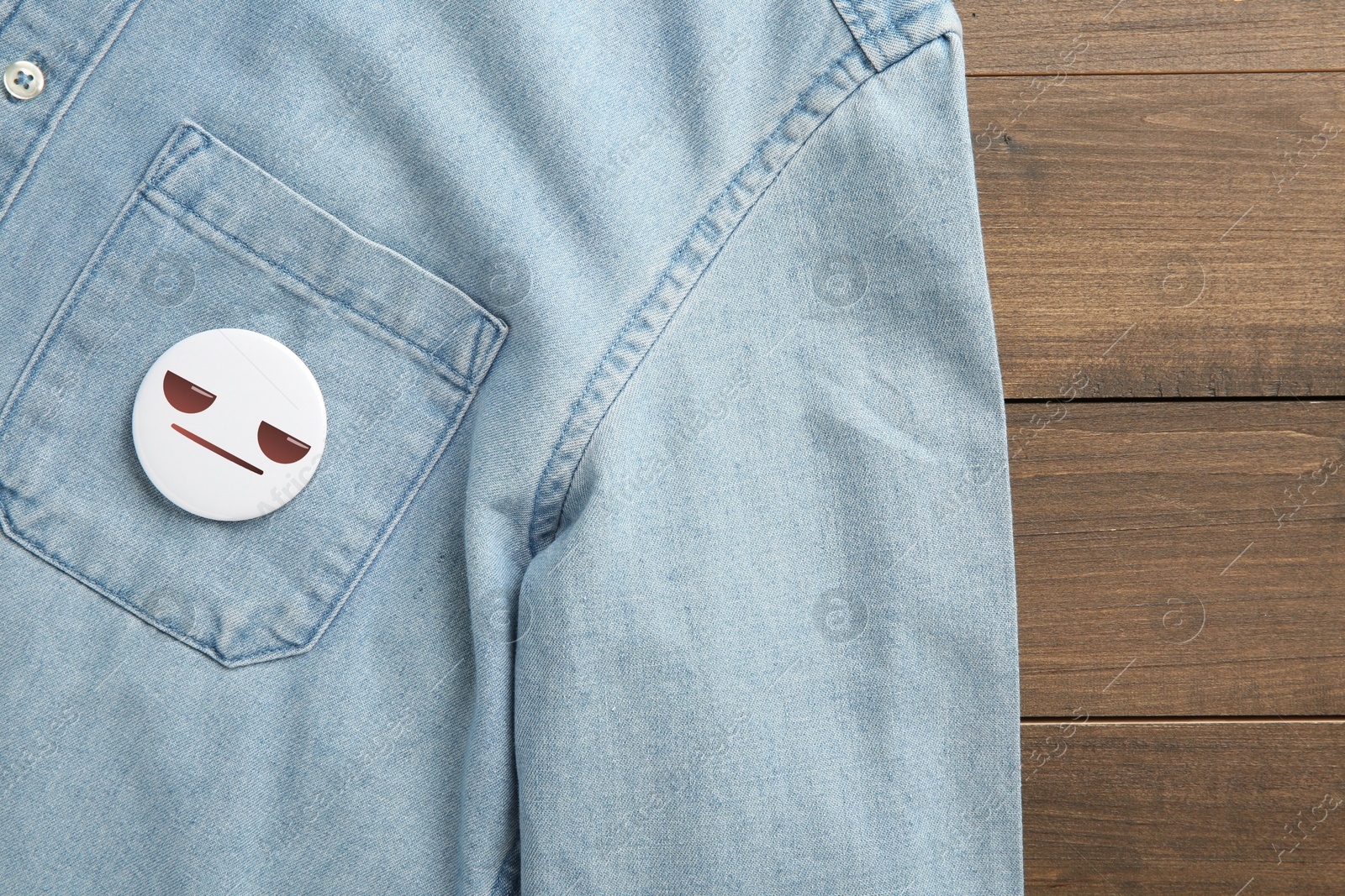 Image of Badge with distrustful or displeased face attached to pocket of denim shirt on wooden background, top view