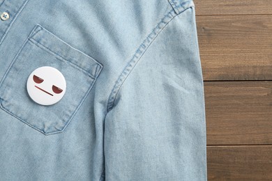 Badge with distrustful or displeased face attached to pocket of denim shirt on wooden background, top view