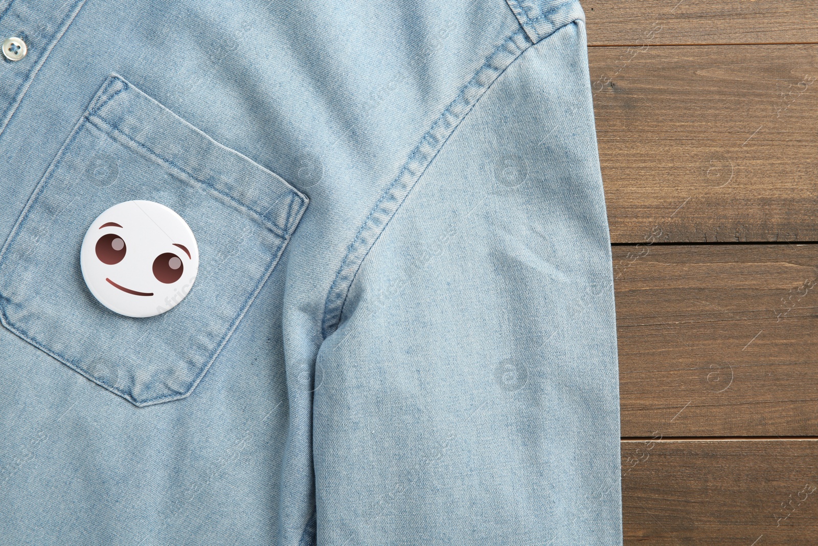 Image of Badge with smiling face attached to pocket of denim shirt on wooden background, top view