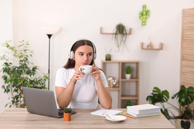 Photo of Student in headphones enjoying hot drink while studying at wooden table indoors