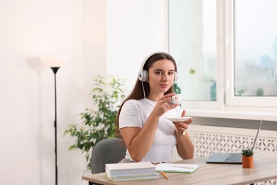 Photo of Student in headphones enjoying hot drink while studying at wooden table indoors