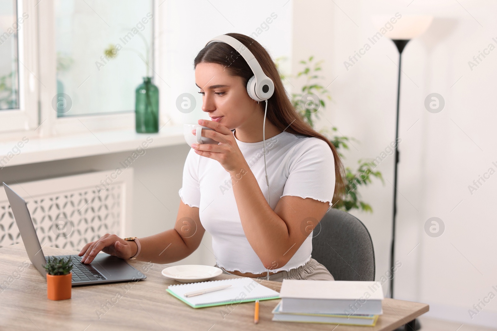 Photo of Student in headphones enjoying hot drink while studying with laptop at wooden table indoors