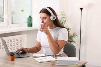 Student in headphones enjoying hot drink while studying with laptop at wooden table indoors