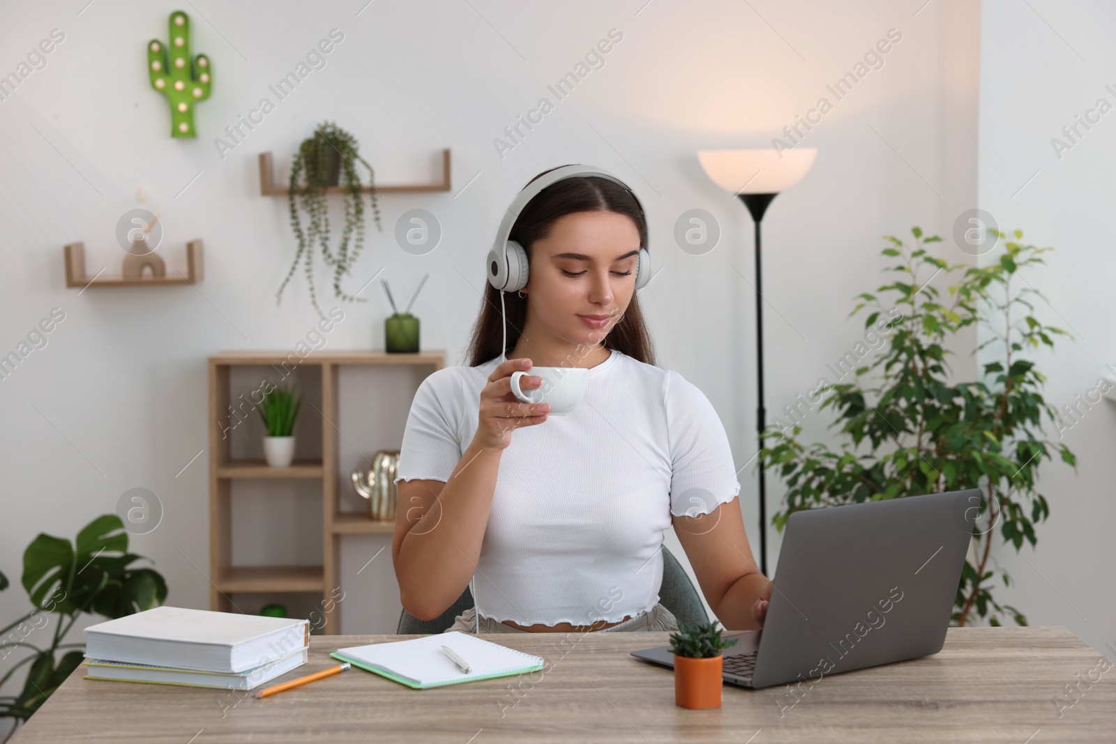 Photo of Student in headphones enjoying hot drink while studying with laptop at wooden table indoors