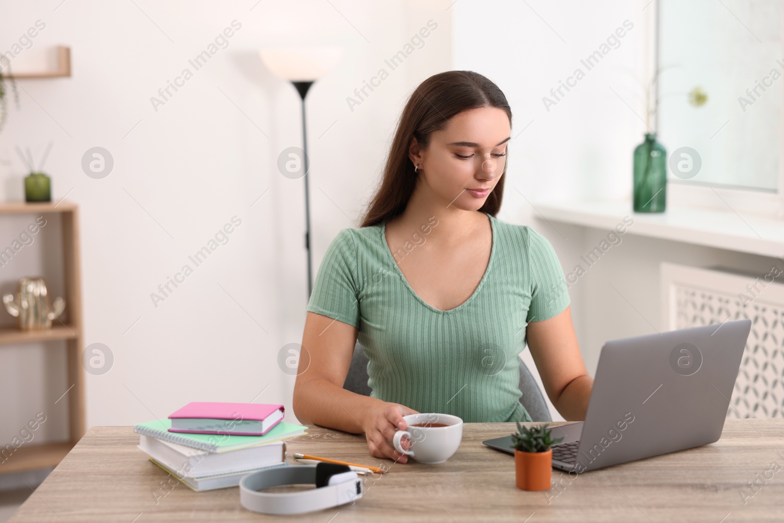 Photo of Student with cup of drink studying at wooden table indoors