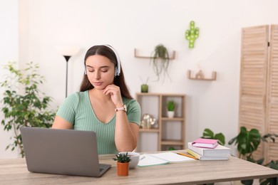 Photo of Student in headphones studying with laptop at wooden table indoors