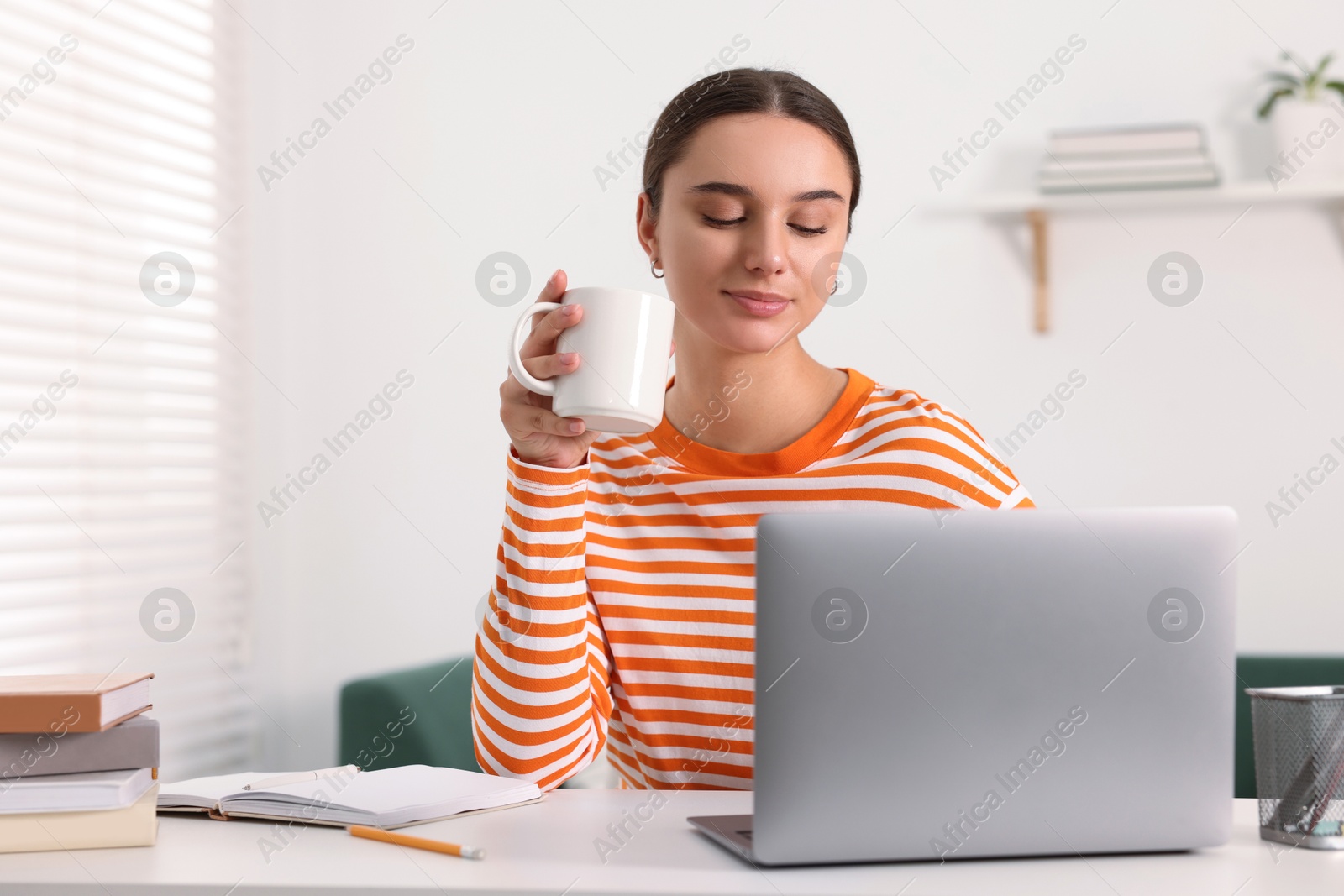 Photo of Student enjoying hot drink while studying with laptop at table indoors