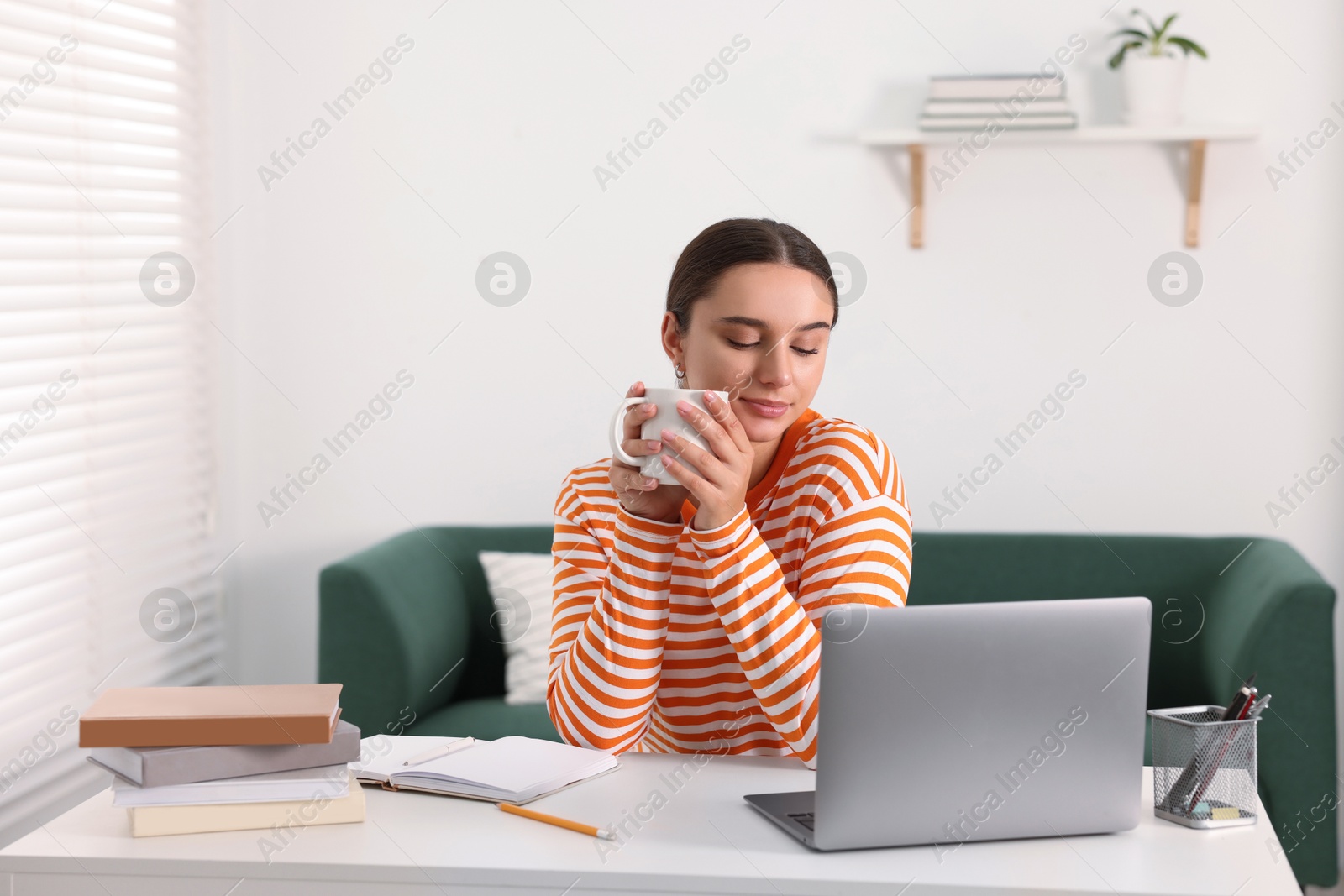 Photo of Student enjoying hot drink while studying with laptop at table indoors