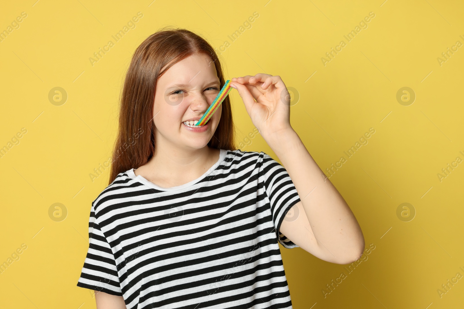 Photo of Teenage girl eating tasty rainbow sour belt on yellow background