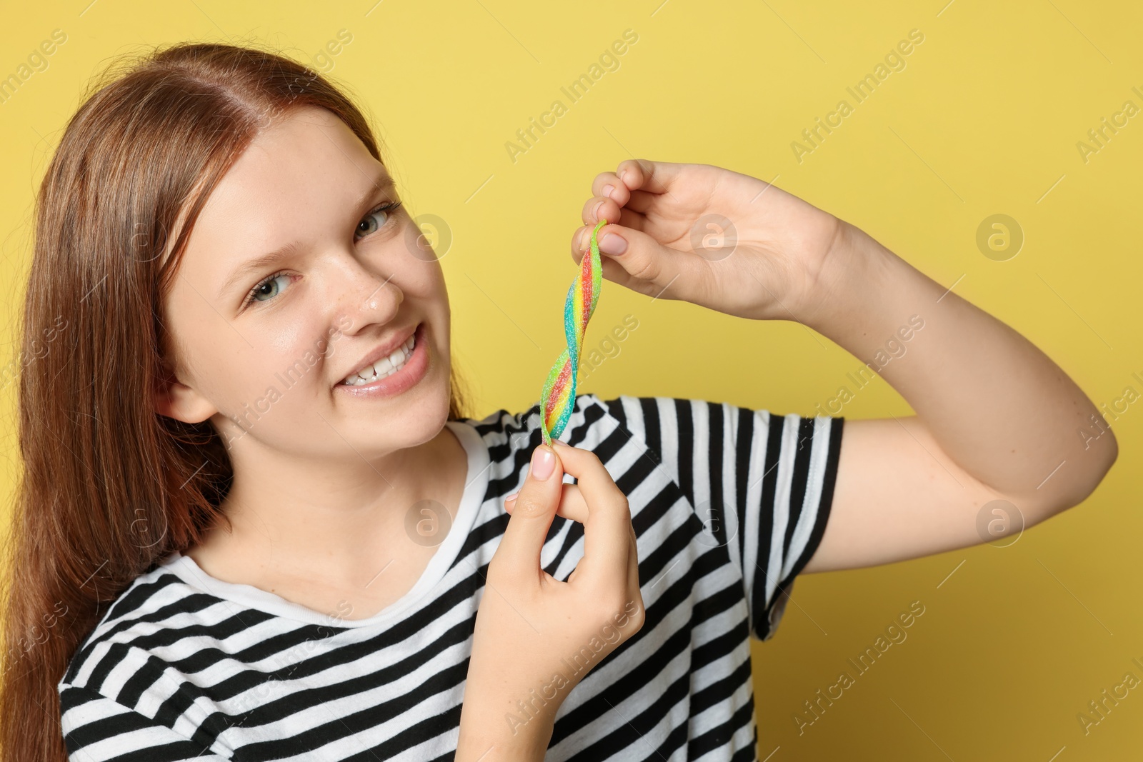 Photo of Happy teenage girl with tasty rainbow sour belt on yellow background