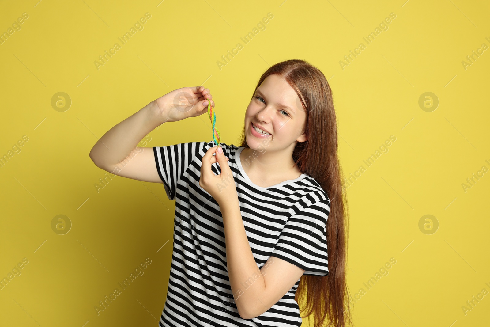 Photo of Happy teenage girl with tasty rainbow sour belt on yellow background