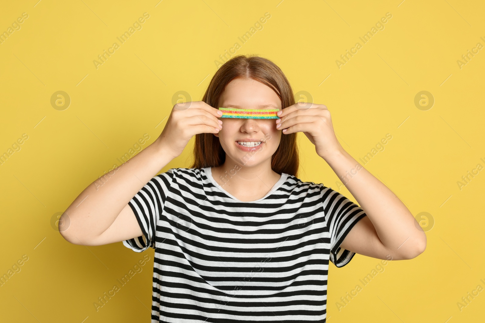 Photo of Happy teenage girl with tasty rainbow sour belt closing her eyes on yellow background