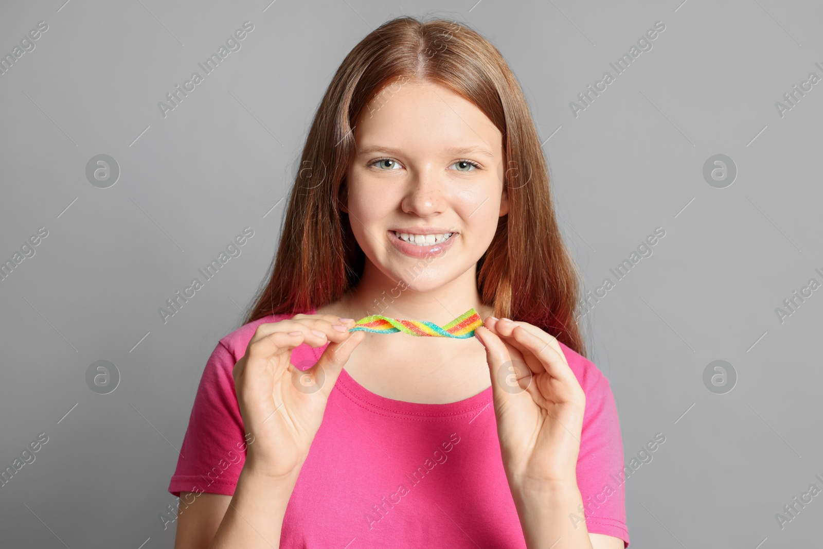 Photo of Happy teenage girl with tasty rainbow sour belt on grey background