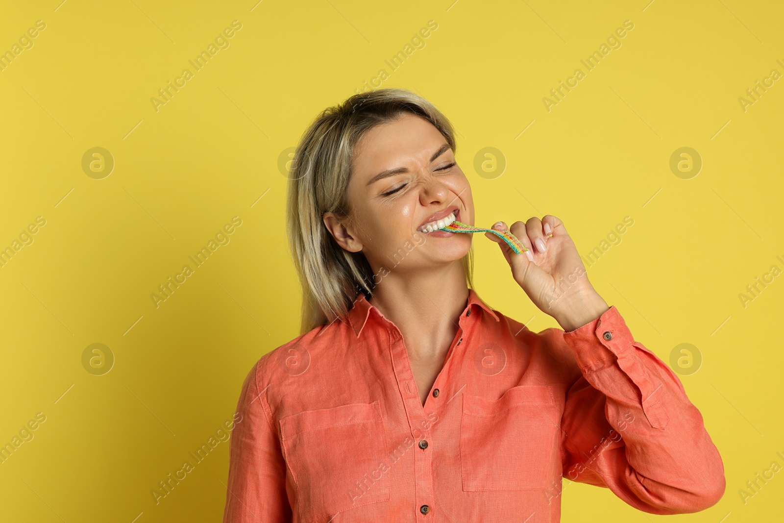 Photo of Young woman eating tasty rainbow sour belt on yellow background