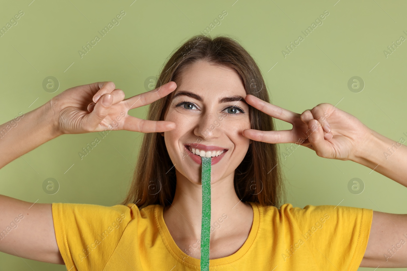 Photo of Happy young woman eating tasty gummy candy while showing v-sign on olive background