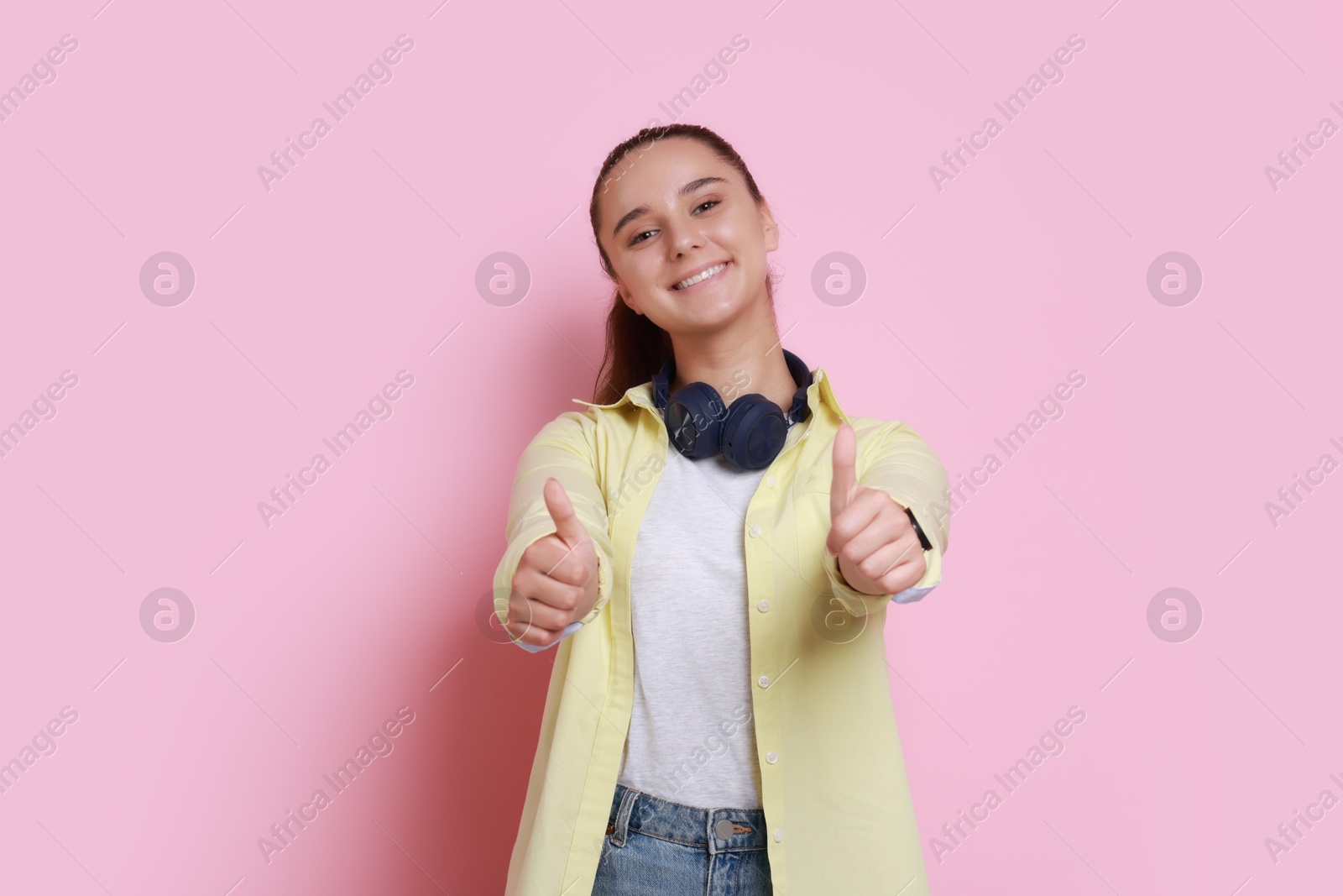 Photo of Happy young student with headphones showing thumbs up on pink background