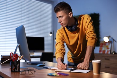 Designer taking notes while working with computer indoors at night