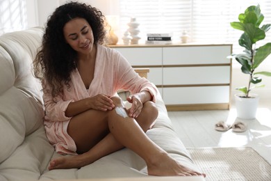 Young woman applying cream onto leg on sofa at home. Space for text