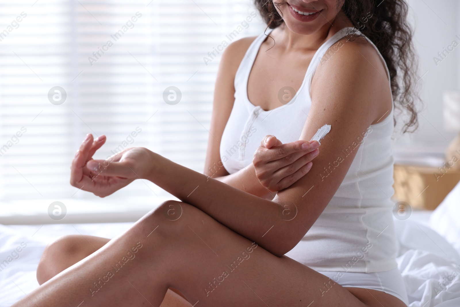 Photo of Woman applying cream onto arm on bed at home, closeup