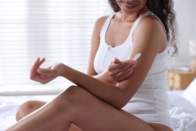 Photo of Woman applying cream onto arm on bed at home, closeup