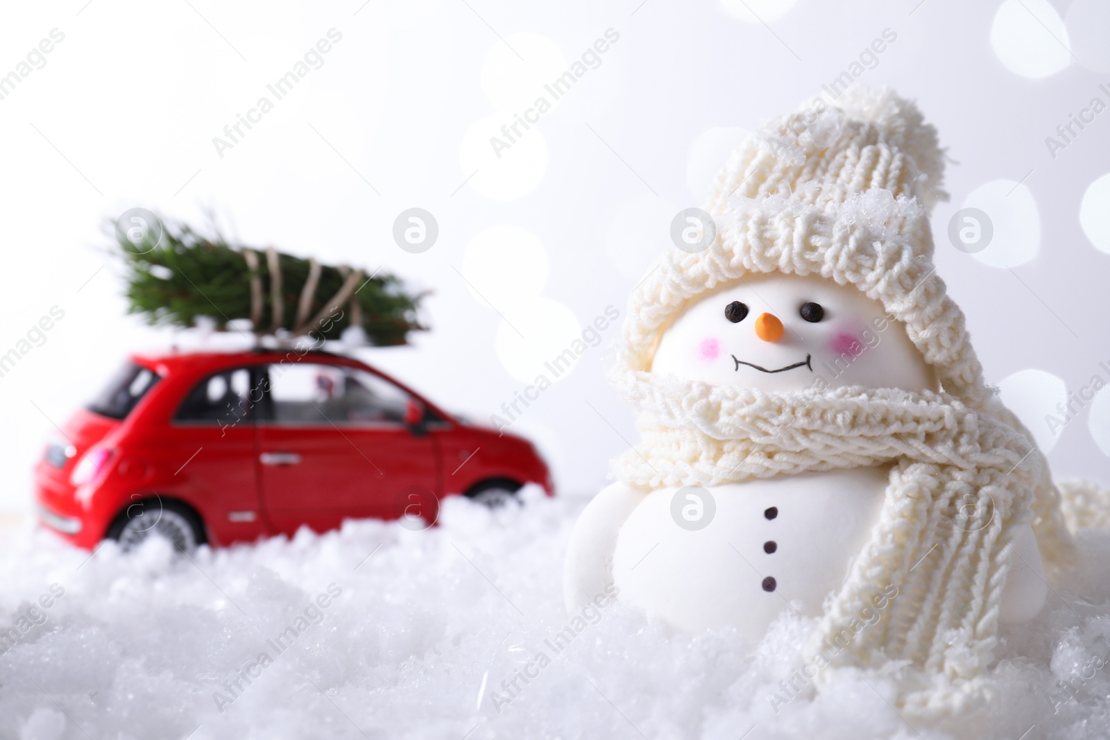 Photo of Cute decorative snowman and toy car with fir tree branches on artificial snow against light background, selective focus