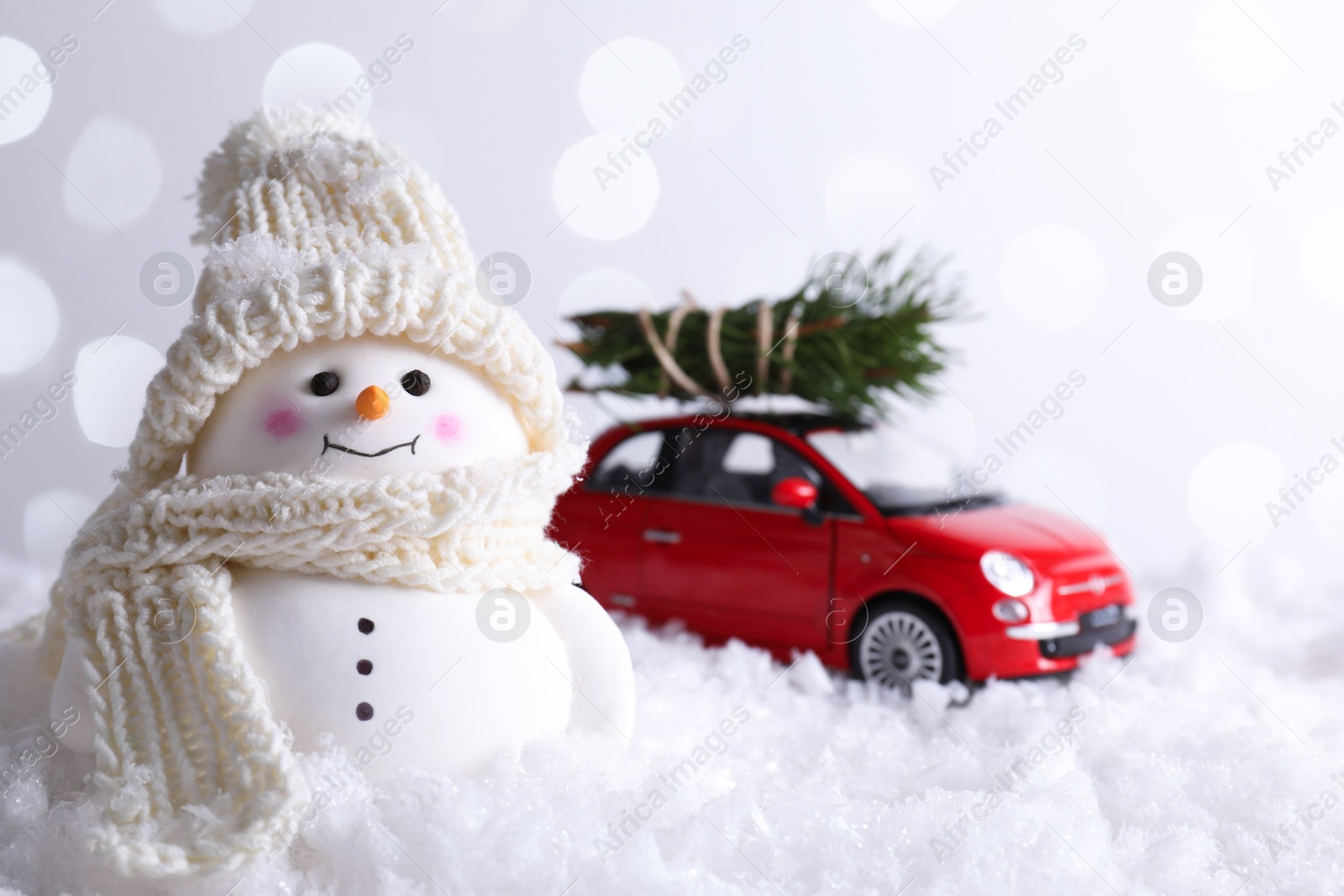 Photo of Cute decorative snowman and toy car with fir tree branches on artificial snow against light background, selective focus