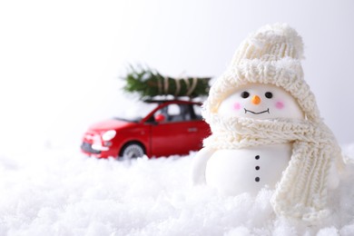 Photo of Cute decorative snowman and toy car with fir tree branches on artificial snow against light background, selective focus