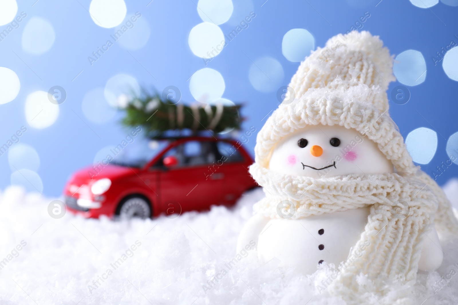 Photo of Cute decorative snowman and toy car with fir tree branches on artificial snow against light blue background, selective focus