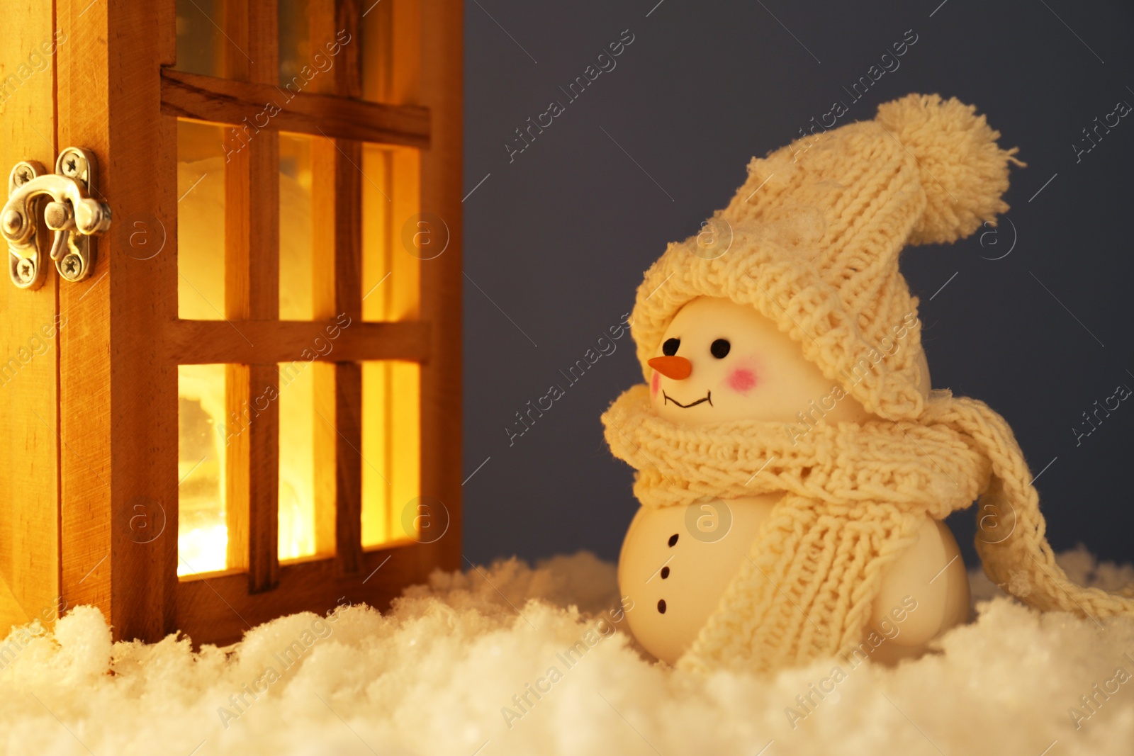 Photo of Cute decorative snowman and wooden lantern on artificial snow against gray background, closeup