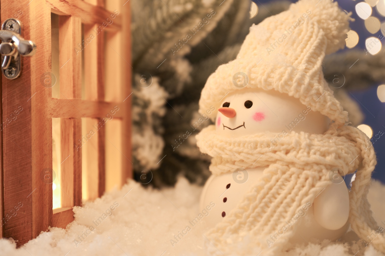 Photo of Cute decorative snowman and wooden lantern on artificial snow against blurred lights, closeup