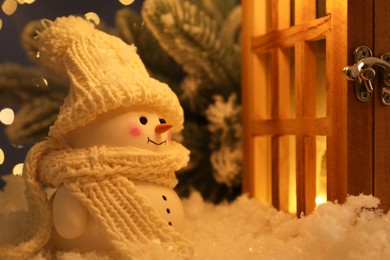 Photo of Cute decorative snowman and wooden lantern on artificial snow against blurred lights, closeup