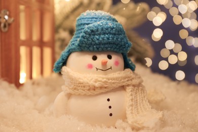 Photo of Cute decorative snowman and other decor on artificial snow against blurred lights, closeup