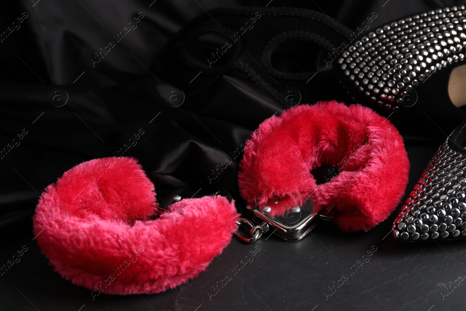 Photo of Red fluffy handcuffs, mask, shoes and fabric on black surface, closeup