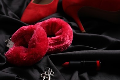 Photo of Red fluffy handcuffs, keys, lipstick and shoes on black fabric, closeup