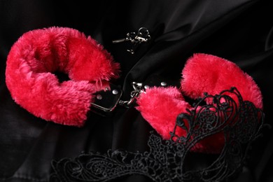 Photo of Red fluffy handcuffs, keys and lace mask on black fabric, closeup