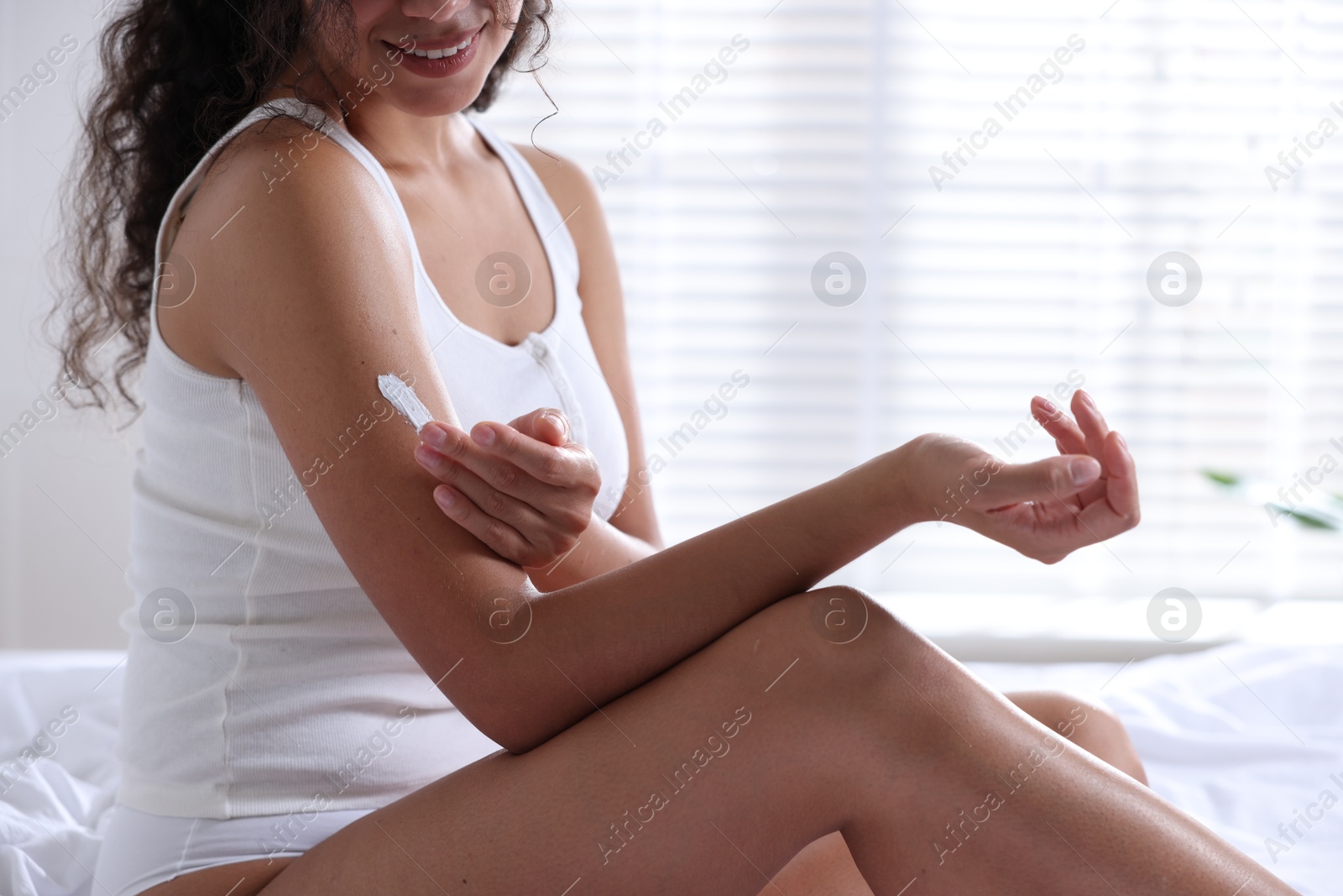 Photo of Woman applying cream onto arm on bed at home, closeup