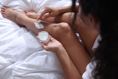 Young woman applying cream onto leg on bed