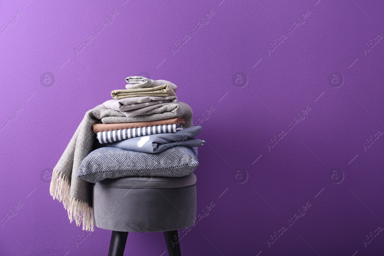 Photo of Stack of clean bed sheets and pillow on stool against purple background. Space for text